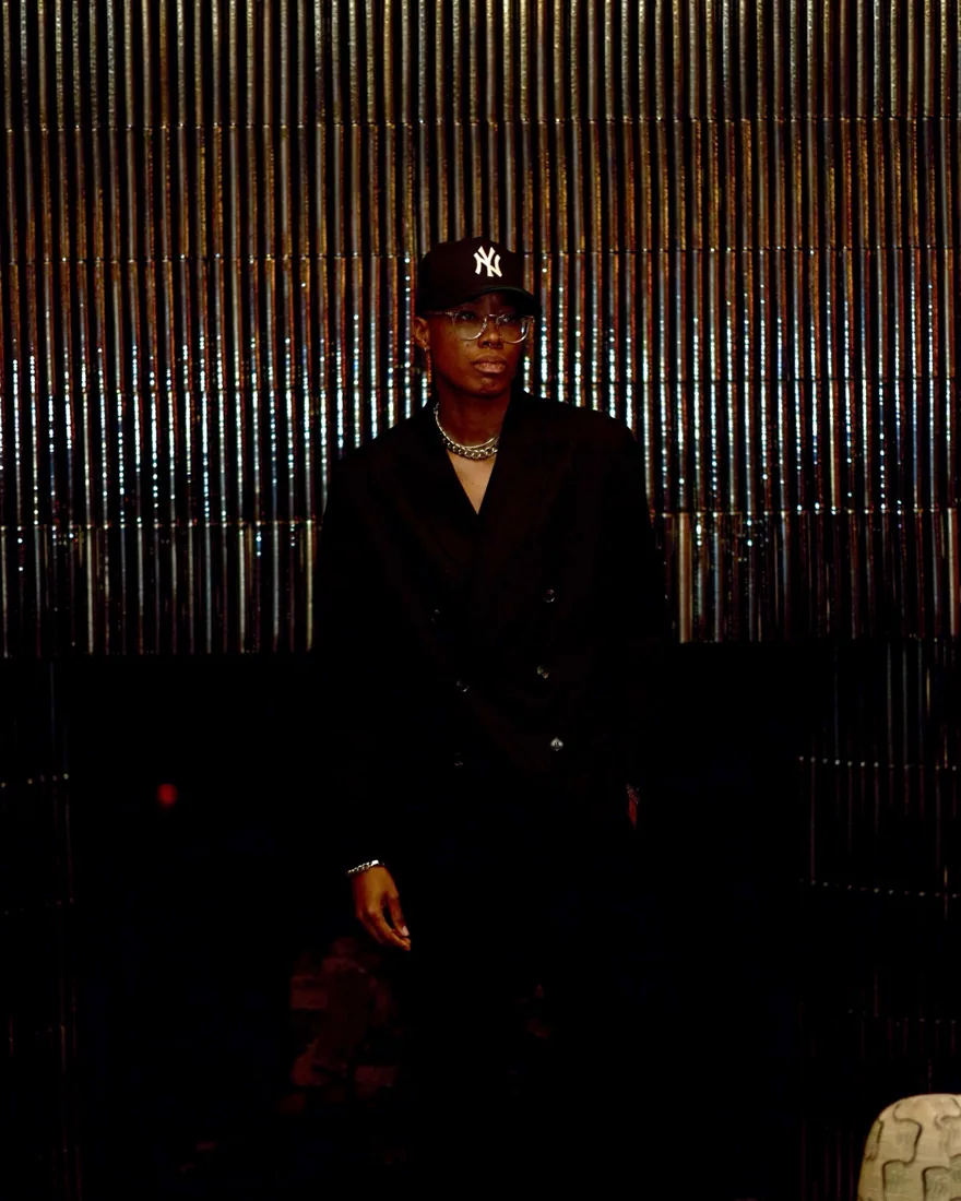 Portrait of Kay Wheeler in a black jacket and cap against a shimmering metallic wall